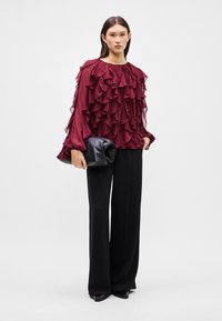 Burgundy ruffled blouse with long sheer sleeves, paired with black wide-leg trousers and a black clutch. Smooth texture, layered design.