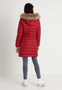 Röd puffjacka med faux fur-huva, mönstrad quiltning och en figurnära silhuett. I kombination med grå jeans och vita högstövlade sneakers.