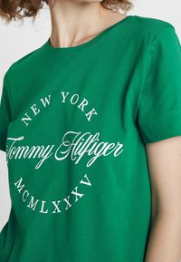 Camiseta de algodón verde con texto impreso en blanco "NUEVA YORK Tommy Hilfiger MCMLXXXV" en disposición curva. Cuello redondo clásico y mangas cortas.