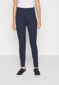 Pantalons ajustés bleu marine avec une taille haute, texture lisse et silhouette élancée, associés à des baskets foncées.