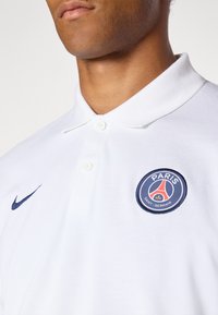 Hvid polo shirt med et blåt Nike-logo og et rundt broderet Paris Saint-Germain badge på venstre bryst. Struktureret stof med krave.