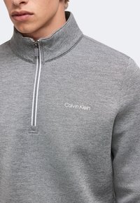 Jeune personne portant un pull Calvin Klein gris à fermeture éclair partiellement fermée et logo sur la poitrine.
