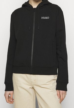 Henkilö, jolla on yllään musta vetoketjullinen huppari, jossa on "HUGO"-logo vasemmassa rinnassa, ja beiget housut, seisoo yksinkertaisen taustan edessä.