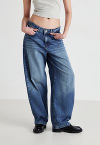 Bredde ben blå jeans i falmet denim med høy livhøyde, subtile slitasjemerker og knappestolpe. Kombinert med svarte loafers.