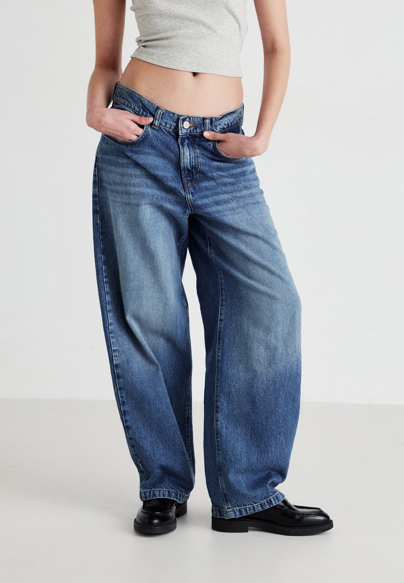 Bredde ben blå jeans i falmet denim med høy livhøyde, subtile slitasjemerker og knappestolpe. Kombinert med svarte loafers.
