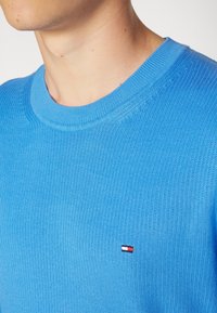 Maglione lavorato a maglia di colore azzurro chiaro con colletto rotondo e polsini a coste, dotato di un piccolo logo rosso, bianco e blu sul petto.