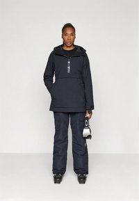 Veste d'hiver bleu marine avec fermeture éclair à l'avant, capuche réglable et pantalon assorti. Comprend des poches cargo et des lunettes de ski à la main. Matériau imperméable.