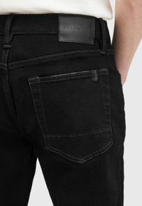 Jeans de mezclilla negra con un parche de cuero en la parte trasera que lleva la inscripción "ALLSAINTS", dos bolsillos traseros y un detalle de costura en contraste.