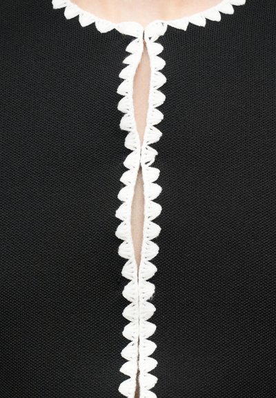 Μαύρο υφασμάτινο με υφή, με κεντρικό κάθετο άνοιγμα περιβαλλόμενο από λευκή κυματοειδή διακόσμηση που αποκαλύπτει το δέρμα κοντά στη γραμμή του λαιμού.