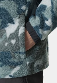 Chaqueta de forro polar con patrón de camuflaje en teal y gris. Cuenta con un bolsillo lateral y puños elásticos para un ajuste ceñido. Textura suave.