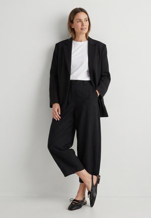 Vrouw staat tegen de muur, draagt een zwarte blazer, wit overhemd, brede zwarte broek en zwarte veterschoenen, kijkt opzij.