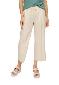 Pantalons beige légers coupés avec une taille élastique et un cordon de serrage. Texture lisse. Associés à des sandales à brides tressées pour le contraste.