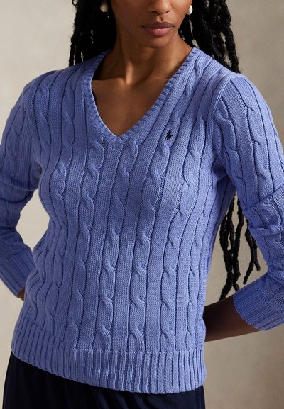 Kobieta w lawendowym swetrze z dzianiny z dekoltem w serek i długimi rękawami oraz ciemnych spodniach, stojąca z rękami na biodrach na jednolitym tle.