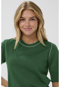 Mladá žena s blond vlasy se usmívá, na sobě má zelený tričko s krátkým rukávem a bílými stehy, na jednobarevném světlém pozadí.