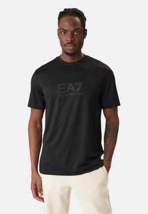 Hombre con cabello trenzado vistiendo una camiseta negra de EA7 Emporio Armani y pantalones de color claro, de pie con una mano en el bolsillo.