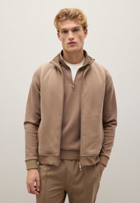 Ärmellose Weste mit Reißverschluss über einem passenden Langarmoberteil, gefertigt aus einem weichen Stoff. Verfügt über einen hohen Kragen und einen glatten Reißverschluss vorne.