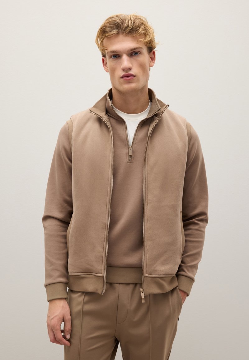 Ärmellose Weste mit Reißverschluss über einem passenden Langarmoberteil, gefertigt aus einem weichen Stoff. Verfügt über einen hohen Kragen und einen glatten Reißverschluss vorne.