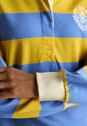 Person iført en langærmet rugbytrøje med brede gule og blå vandrette striber og hvide manchetter, armene krydset ved taljen.