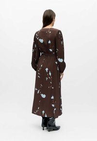 Mujer de espaldas, vestida con un vestido largo marrón con patrones florales azul claro, mangas largas, cinturón en la cintura y botas negras de tacón.