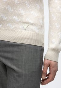 Μπεζ πουλόβερ με σχέδια και λογότυπο GUESS πάνω από ριμπ τελείωμα, φορεμένο με γκρι παντελόνι με υφή και χαλαρό δεξί χέρι εμφανές.