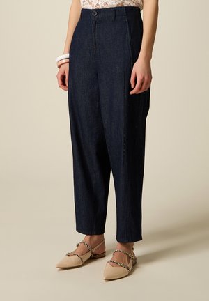 Proste spodnie o prostych nogawkach w ciemnym niebieskim kolorze, noszone z beżowymi, spiczastymi balerinami z cienkimi paskami i bransoletkami na kostkę, widoczne od talii do stóp.