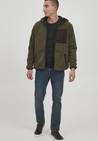 Olive green, fuzzy jacket with a black zippered chest pocket, front pockets, and adjustable hood, paired with dark jeans and black sneakers.