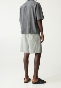 Chemise grise à manches courtes, coupe décontractée, associée à un short vert clair avec une poche arrière, et des sandales noires à brides ajustables.