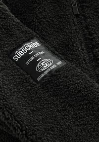 Veste en polaire noire avec une surface texturée, arborant une étiquette rectangulaire sur laquelle est inscrit "ABONNEZ-VOUS," "SYSTÈMES DU FUTUR," et "ÉTABLI DEPUIS 1982."