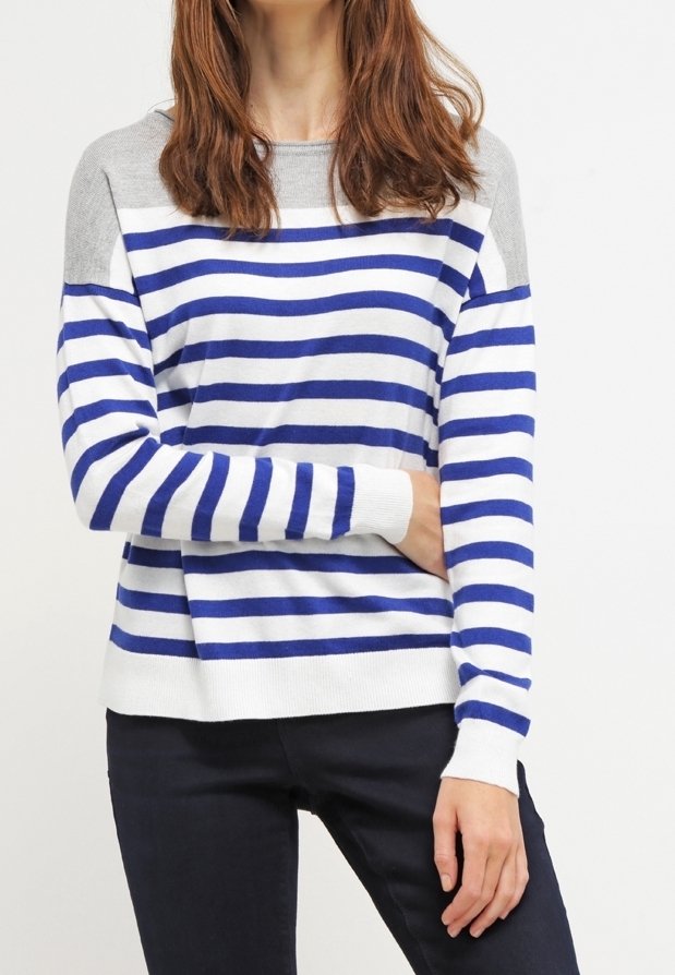 Femme portant un pull à manches longues rayé bleu et blanc avec des épaules grises et un pantalon noir, debout devant un fond blanc.