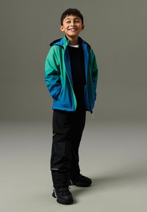 Hymyilevä poika seisoo käsineet taskuissa, yllään vihreä ja sininen takki, mustat housut ja mustat kengät yksiväristä harmaata taustaa vasten.