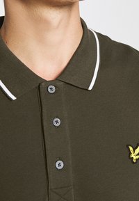 Olivově zelené polo tričko s bílým límcem a dvěma knoflíky. Má texturovanou tkaninu a žluté logo vyšité na hrudi.
