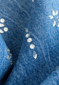 Țesătură de denim albastru cu aplicatii clare, asemănătoare cu pietre prețioase, aranjate în forme florale pe întreaga suprafață, cu textura țesăturii vizibilă.