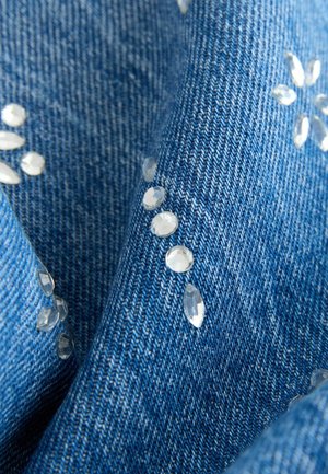 Mėlynos spalvos džinsinis audinys, kuriame aiškiai matomi brangakmenius primenantys papuošimai, išdėstyti gėlės formomis ant paviršiaus, matomas audinio pynimas.