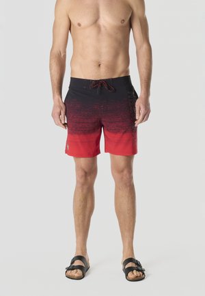 Hombre sin camiseta con pantalones de baño de gradiente rojo y negro y sandalias negras, de pie frente a un fondo claro uniforme.