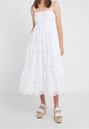Femme portant une robe midi blanche sans manches avec des motifs floraux découpés et des sandales compensées blanches à brides à la cheville.