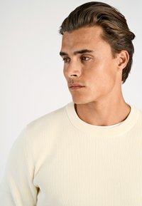 Helle cremefarbene gerippte Strickjacke mit Rundhalsausschnitt, die eine strukturierte Oberfläche und lange Ärmel hat, getragen über einem weißen T-Shirt.