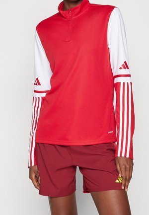 Sportowiec ubrany w czerwoną i białą sportową bluzkę z długim rękawem z paskami na rękawach oraz bordowe szorty z żółtym logo.