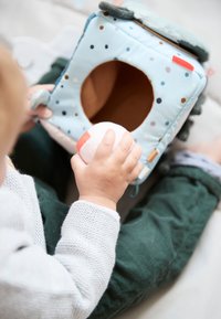 Soft fabric toy cube in light blue with colorful polka dots, a brown interior, and a front hole; child holds a plush ball with a red stripe.