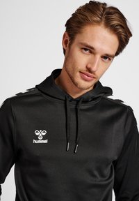 Svart hoodie med vit logotyp och detaljer. Har dragsko och en strukturerad textil. Sidofält ger visuell variation.