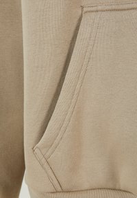 Beige Kapuzenpullover mit vorderer Tasche, gerippten Bündchen und weicher Textur. Der Stoff wirkt glatt mit feinen Nähten entlang der Kanten.