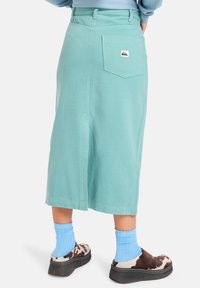 Een middellange, lichtteal denim rok met een achterzak met een logo-patch. Gecombineerd met blauwe sokken en gestreepte instappers.