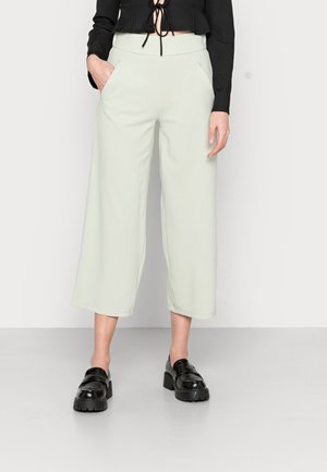 Woman wearing light green wide-leg cropped pants, black long-sleeve crop top, and black chunky loafers, standing against a plain background.