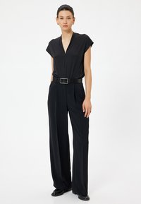 Sort kortærmet jumpsuit med V-halsudskæring, belagt talje med en sølvspænde og vide bukser, lavet af glat stof.