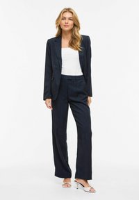 Femme debout, portant un costume noir à fines rayures avec un pantalon à jambes larges, un haut blanc et des talons ouverts de couleur claire, sur fond blanc.