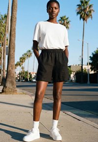 Persoon staat op een zonovergoten straat met palmbomen, draagt een wit t-shirt, zwarte korte broek, witte sokken en witte sneakers, met handen in de zakken.