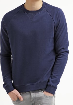 Mann trägt einen einfarbigen dunkelblauen Sweatshirt mit langen Ärmeln, gerippten Bündchen und Saum, Hände teilweise in den Taschen einer hellgrauen Jeans.