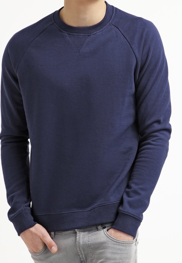 Homme portant un sweat-shirt bleu foncé uni à manches longues avec poignets et ourlet côtelés, mains partiellement dans les poches d'un jean gris clair.