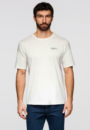 Bärtiger Mann mit dunklem Haar, der ein weißes T-Shirt mit dem Schriftzug "Chase Your Dreams, Not People" trägt, kombiniert mit blauer Jeans vor einem einfarbigen Hintergrund.