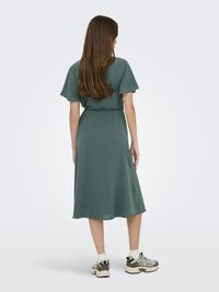 Vestito midi verde con maniche a farfalla corte, vita cintata e orlo fluente. Indossato con sneakers casual e calzini alla caviglia, visto da dietro.