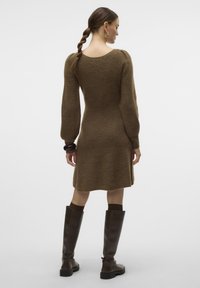 Abito in maglia marrone con corpetto aderente, orlo svasato e maniche lunghe a sbuffo. Indossato con stivali alti fino al ginocchio in pelle marrone. Texture liscia.